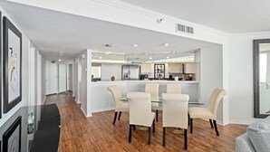 A modern kitchen with a central island, a dining area with a glass table and chairs, and a hallway with a framed artwork. - Oceanfront 3BR Balcony Indoor Outdoor Pools (Daytona Beach Shores)