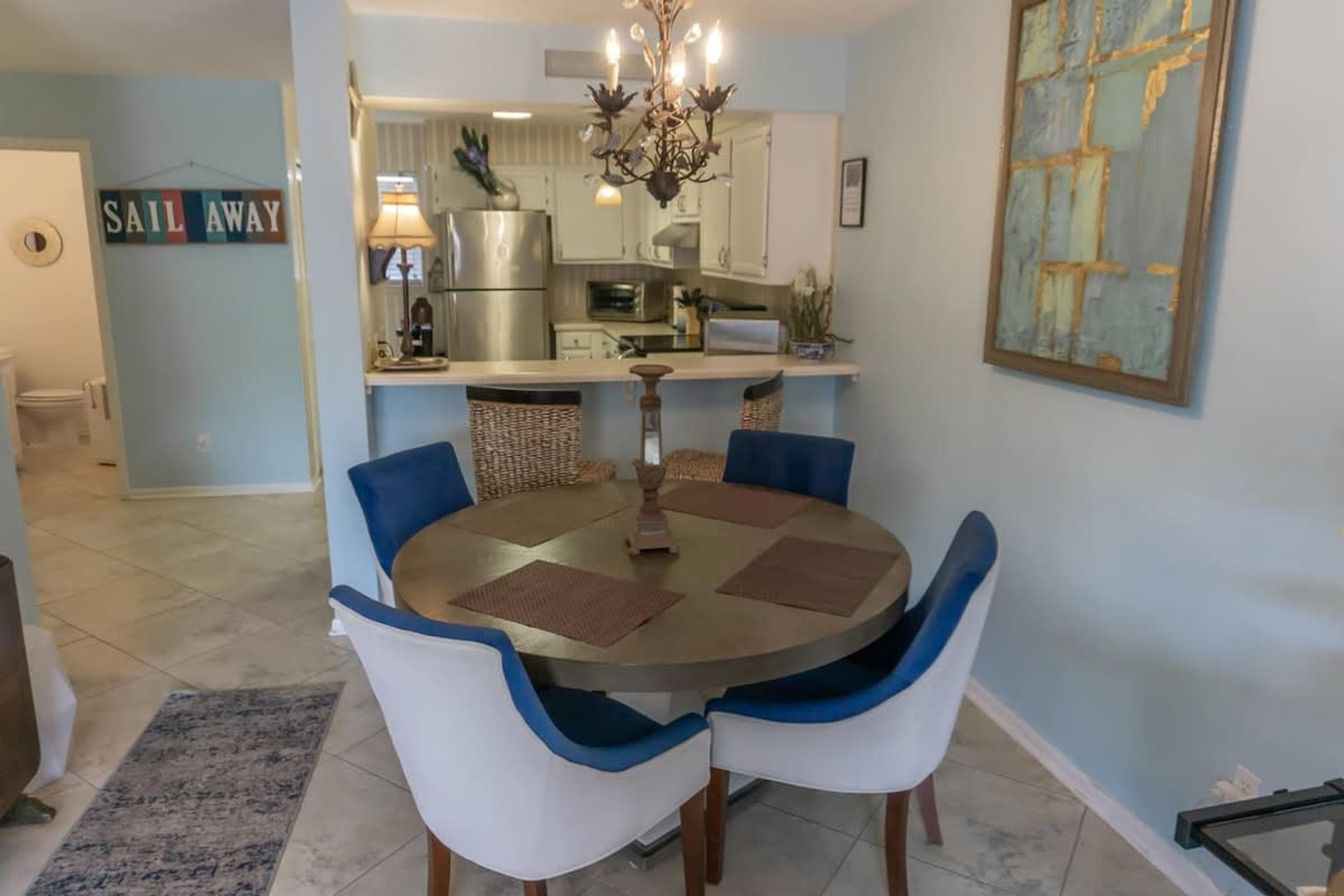 A dining area with a round table, four chairs, a chandelier, and a kitchen in the background.