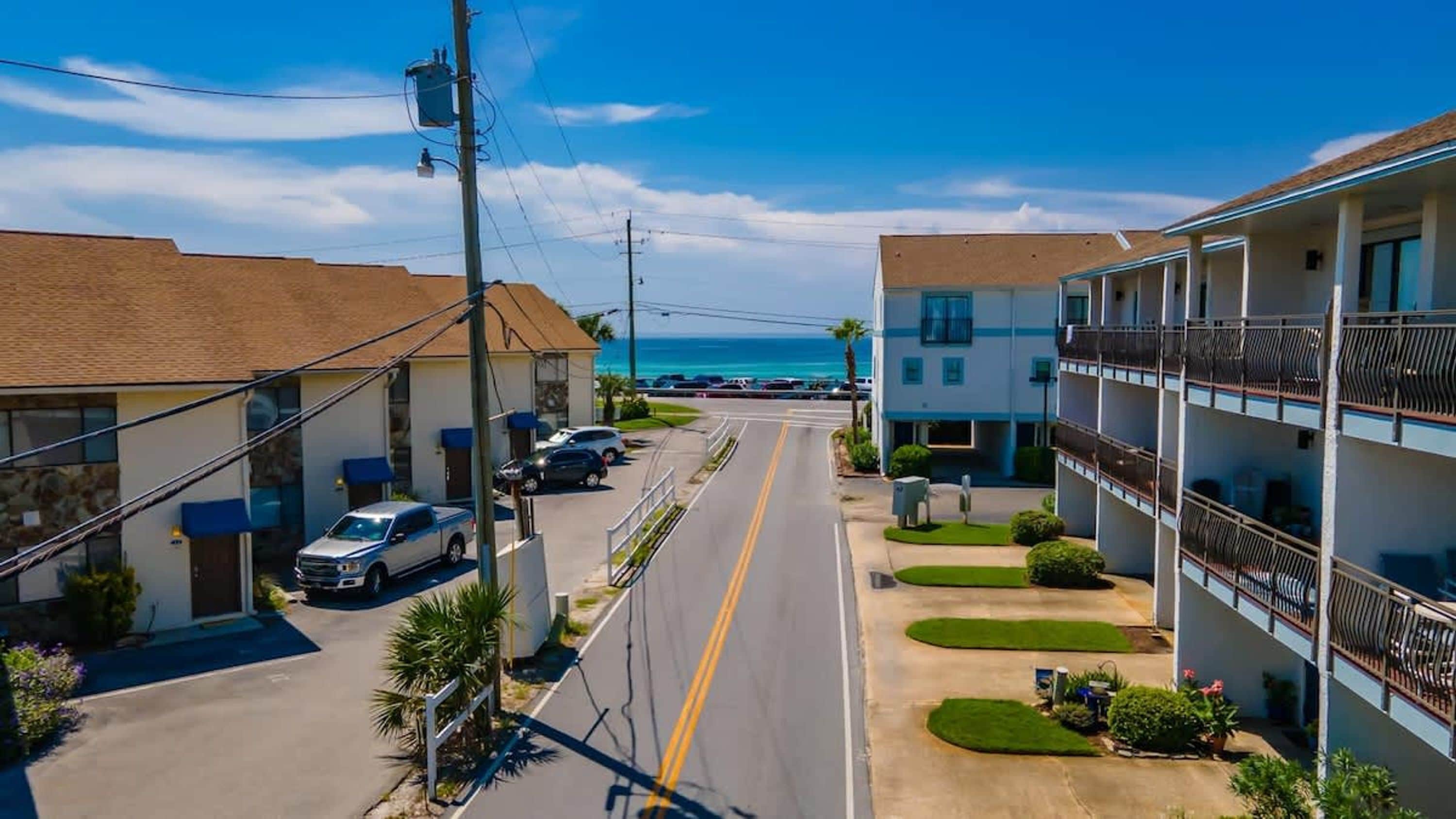 Un hotel frente al mar, con un camino que lleva a la playa, varios balcones y coches aparcados.