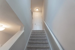 Una escalera con alfombra que lleva a una puerta blanca en la parte superior, una lámpara a la izquierda y un pasamanos blanco a la derecha.