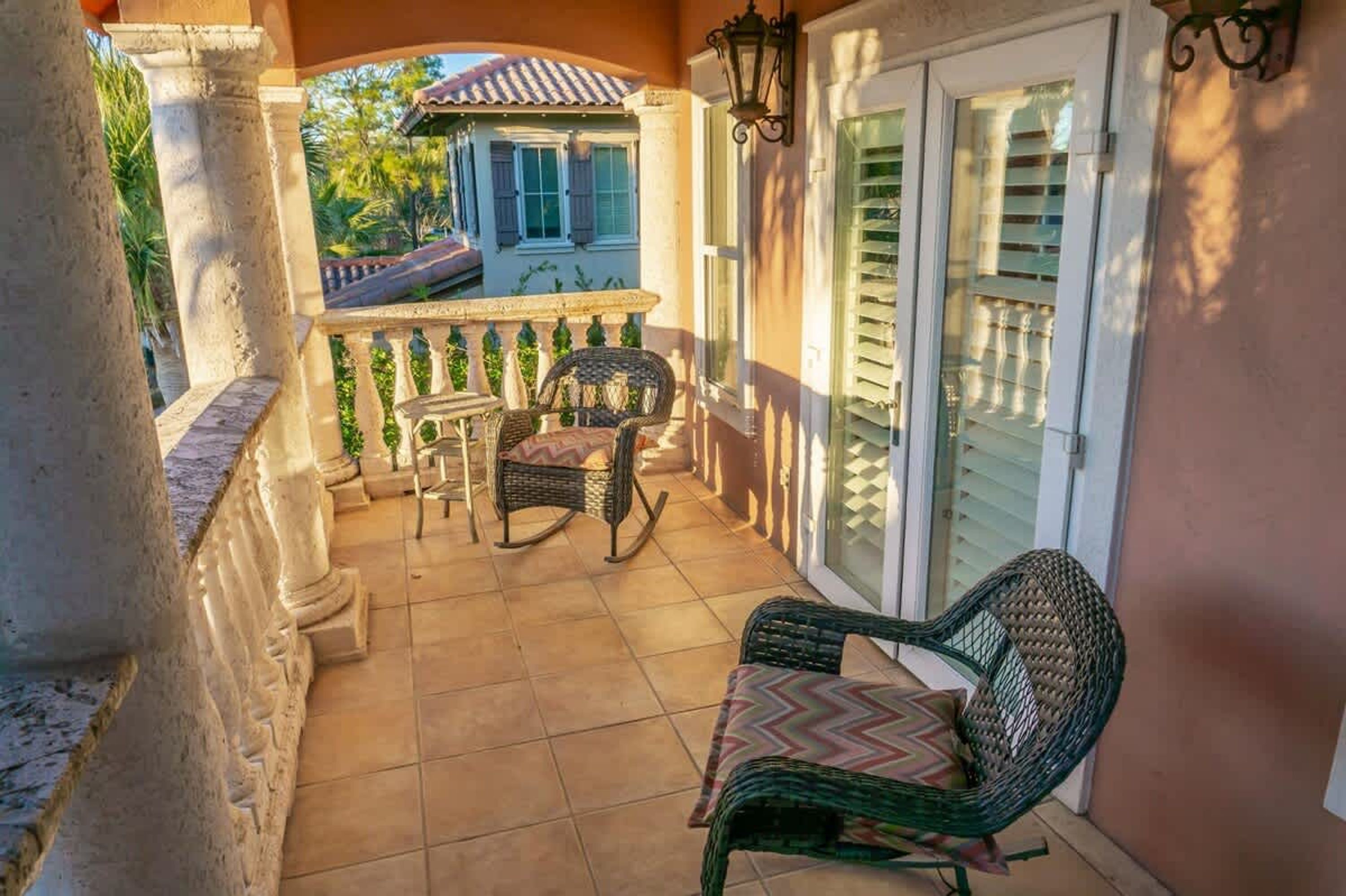 A balcony with a table and chairs, a view of a house, and a door with shutters.