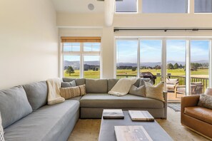 A spacious living room with a large gray sectional sofa, a coffee table, and a leather armchair. The room has large windows offering a view of a golf course and distant hills.