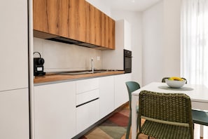 A compact kitchen with white cabinets, a wooden upper section, a black oven, and a dining area with a white table and a green chair. - Elegantis Luce di Trastevere Apartment with Terrace (Roma)