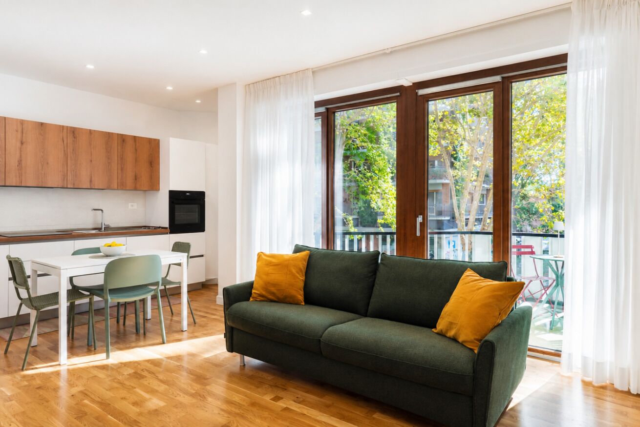 A modern living room with a green sofa, a dining table with chairs, and a kitchen area with wooden cabinets and a black appliance.