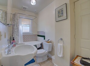 Een badkamer met een bad, toilet en wastafel. Er is een raam met zonwering, een douchegordijn en een lijst met een foto aan de muur.