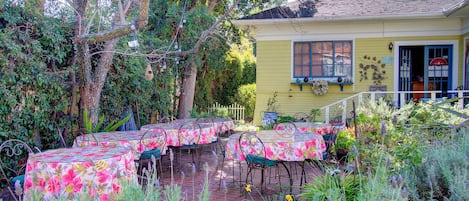 An outdoor dining area with tables covered in floral tablecloths, surrounded by greenery and a yellow building with a window.
