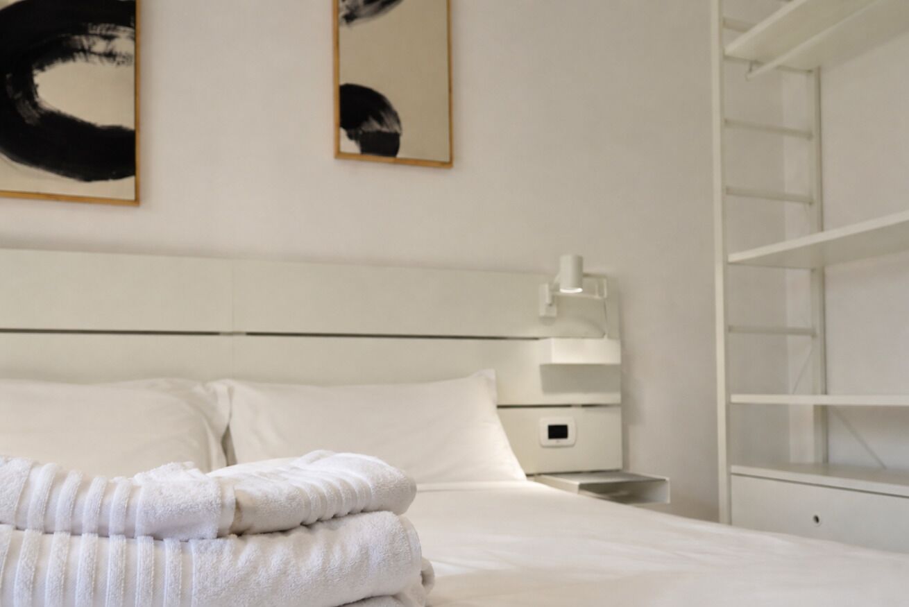 Ein Hotelzimmer mit einem ordentlich bezogenen Bett, einem Nachttisch, einer Lampe und einem Kleiderschrank.