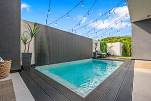 A swimming pool with a tiled edge, surrounded by a wooden deck. There are string lights overhead and a wall with vertical lines. - Hooiberg 49 U Apt 1 Pearl House (Oranjestad)