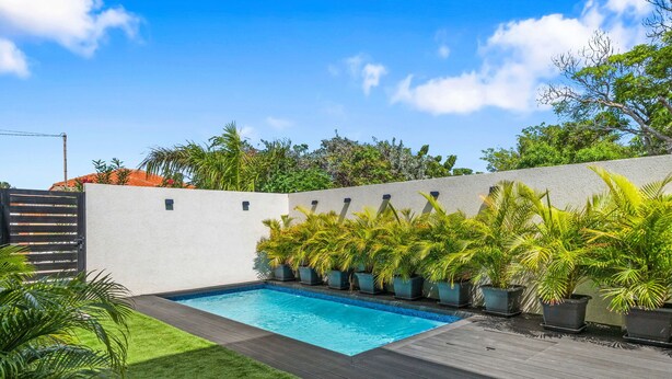 Ein Schwimmbecken mit einer umlaufenden Terrasse, Topfpflanzen und blauem Himmel.