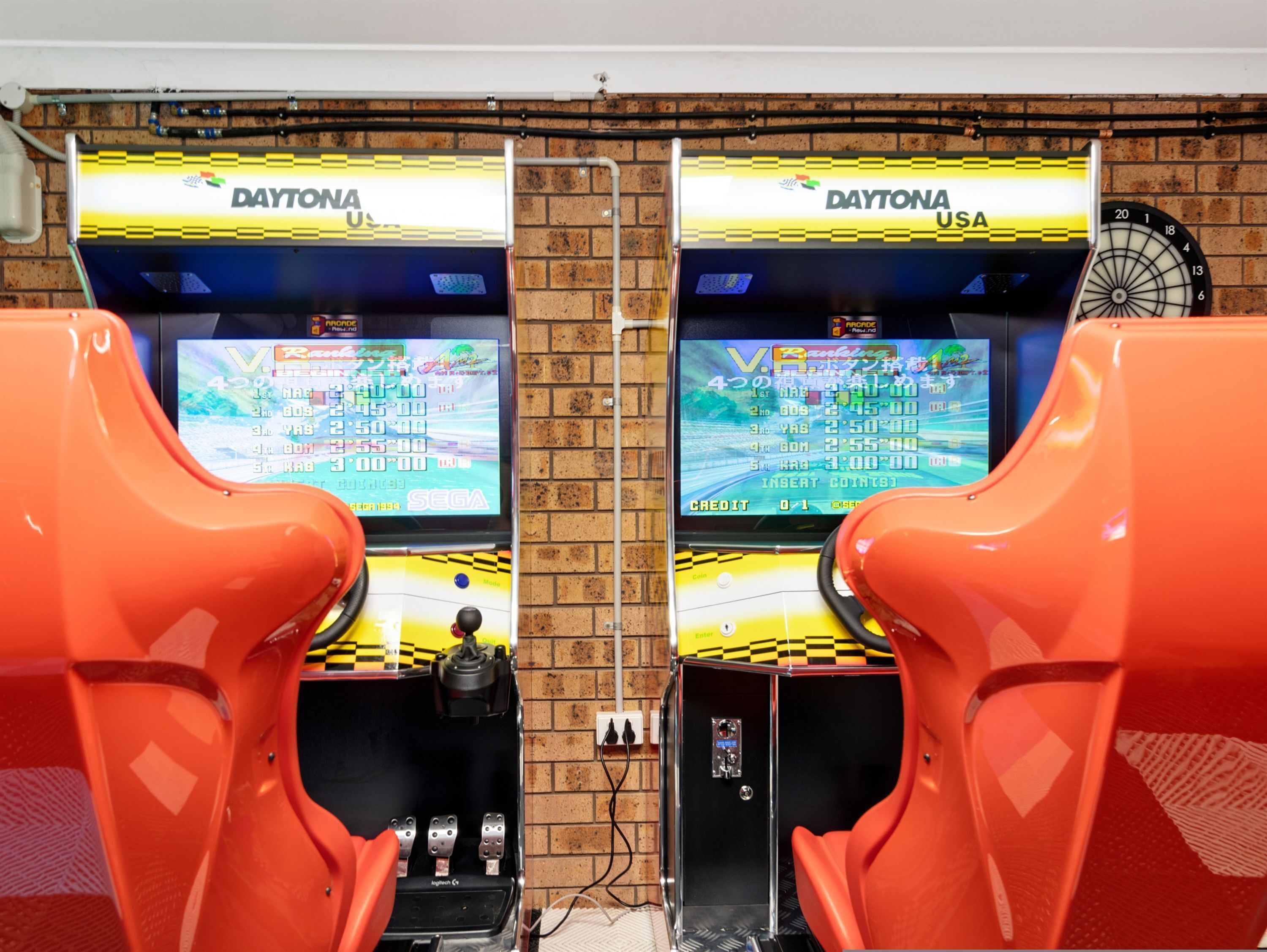 Two arcade racing game machines with a dartboard and a ladder against a brick wall.