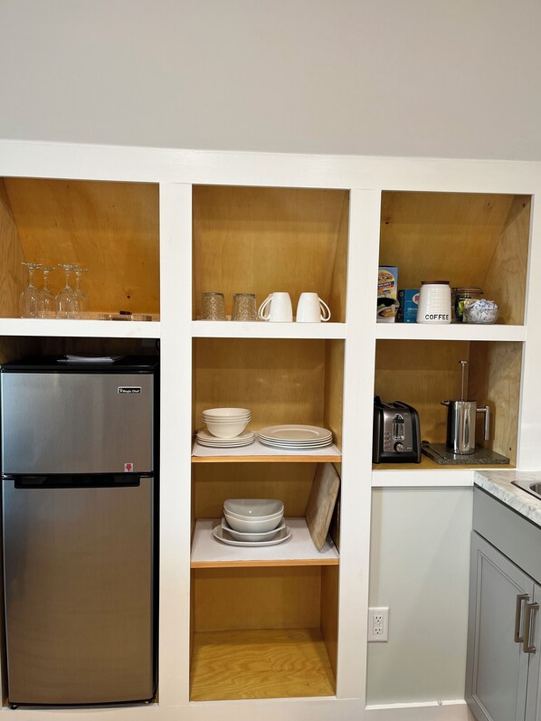 A kitchen with a stainless steel refrigerator, shelves with dishes, glasses, and a coffee maker.