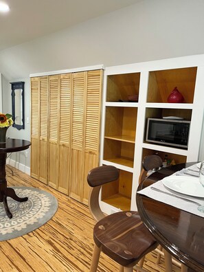 A dining area with a round table, chairs, a wooden floor, a cabinet with shelves, and a microwave.
