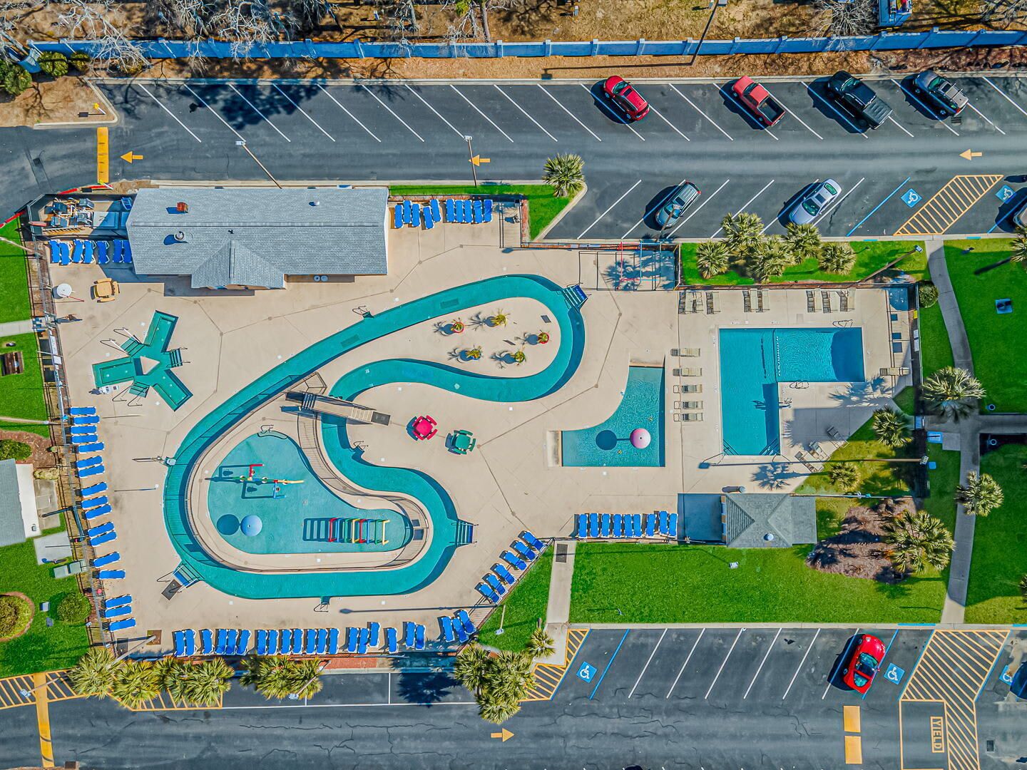 An aerial view of a parking lot with multiple rows of cars, a swimming pool area with a winding water feature, and a building with a sloped roof.
