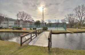 Eine Holzbrücke über ruhigem Wasser, umgeben von Gras und Bäumen, mit Gebäuden im Hintergrund.