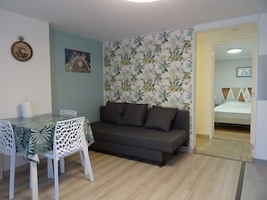 A room with a dining table, white chairs, a sofa, a patterned wallpaper, and a bed in the adjacent room. - Charming, detached garden-level vacation rental near the town center (Neuvic)