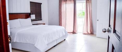 A hotel room with a bed, a bedside table, a window with curtains, and a door.