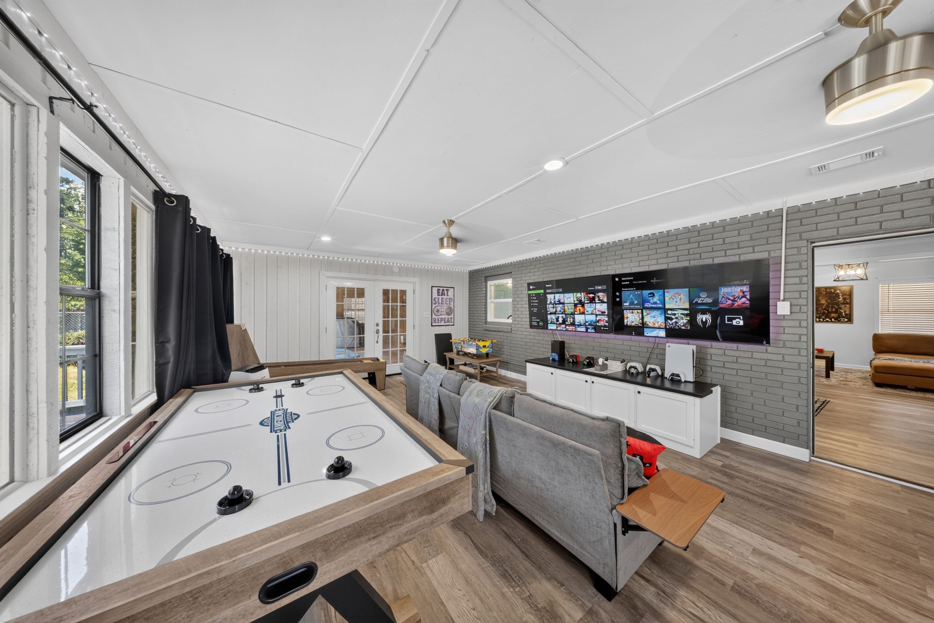 Ein modernes Wohnzimmer mit Shuffleboard-Tisch, grauem Sofa, Spielkonsole und TV-Wand.