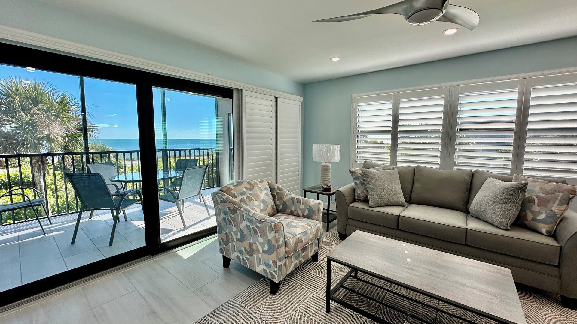 A living room with a sofa, armchair, and coffee table, a balcony with chairs, and a view of the ocean.
