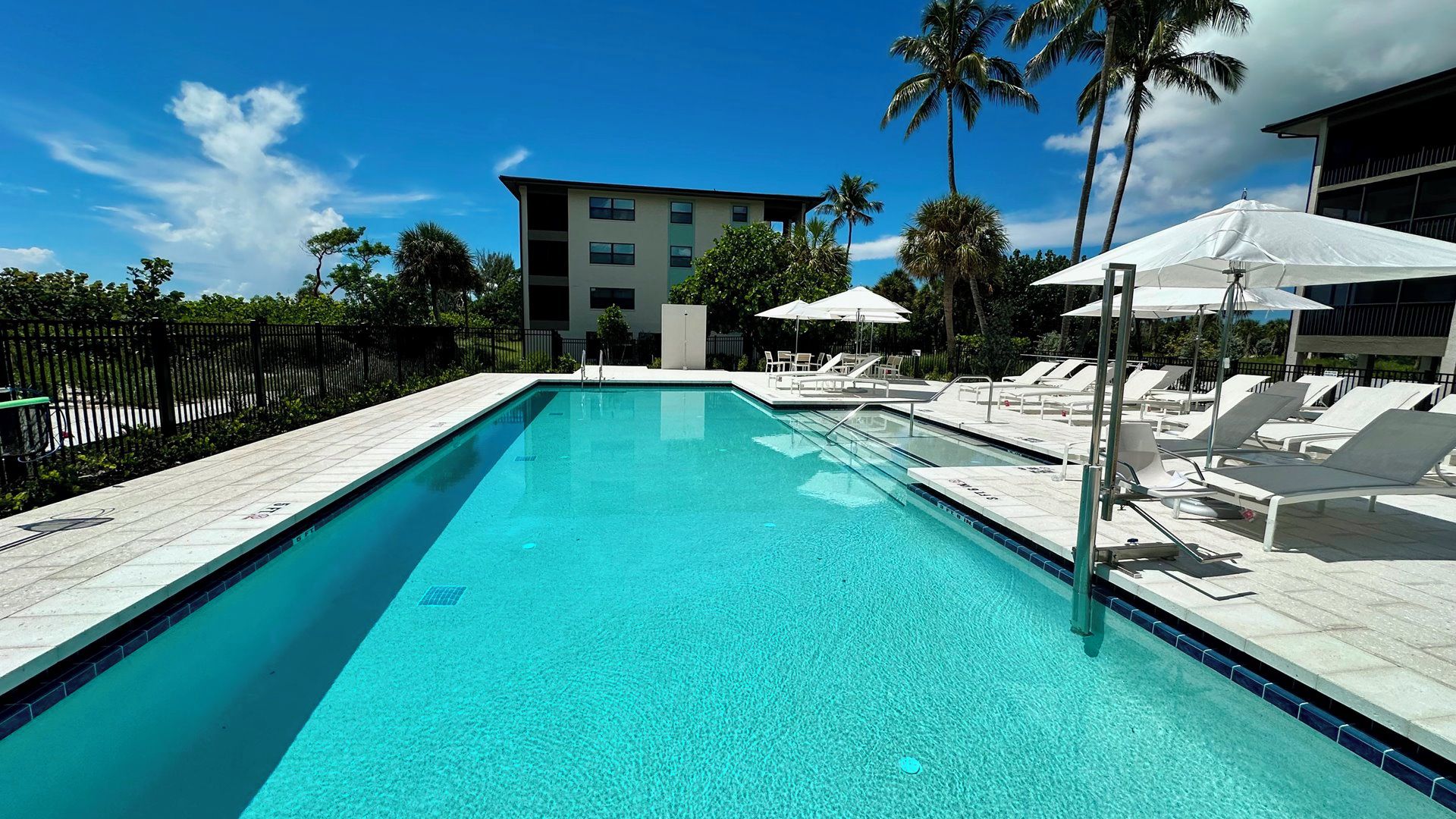 A clear blue pool with lounge chairs and umbrellas, surrounded by palm trees and a building in the background.
