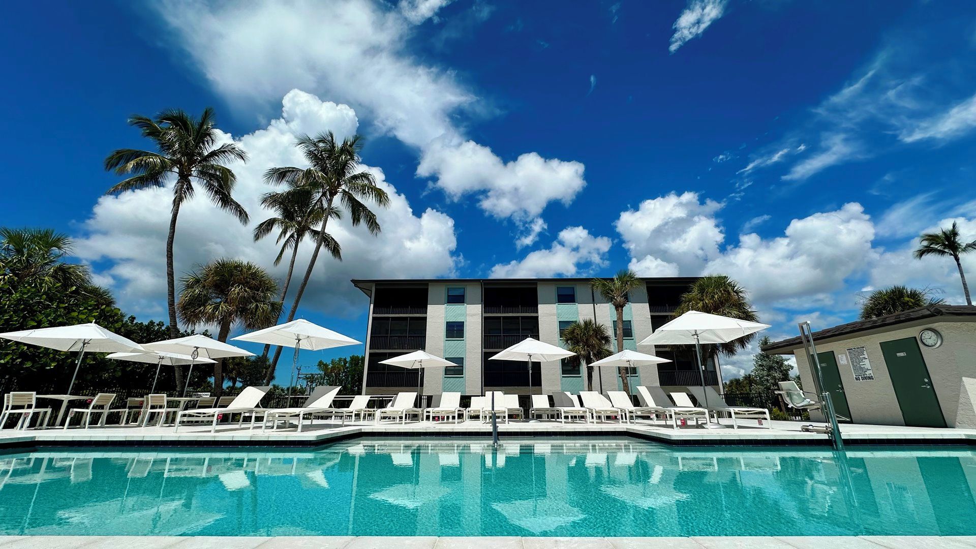 Ein Poolbereich mit Liegestühlen und Sonnenschirmen, ein Gebäude mit vielen Fenstern und Palmen unter blauem Himmel.