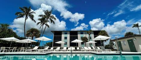 A pool area with lounge chairs and umbrellas, a building with multiple windows, and palm trees under a blue sky.
