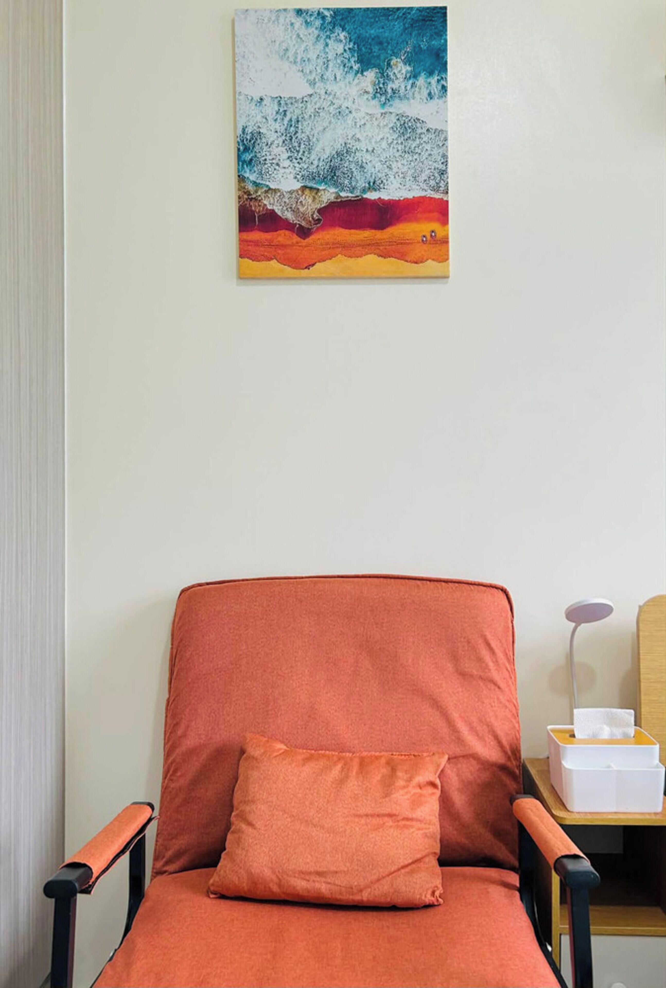 A room with a single orange chair, a cushion, a small table with a tissue box, and a painting of a wave on the wall.