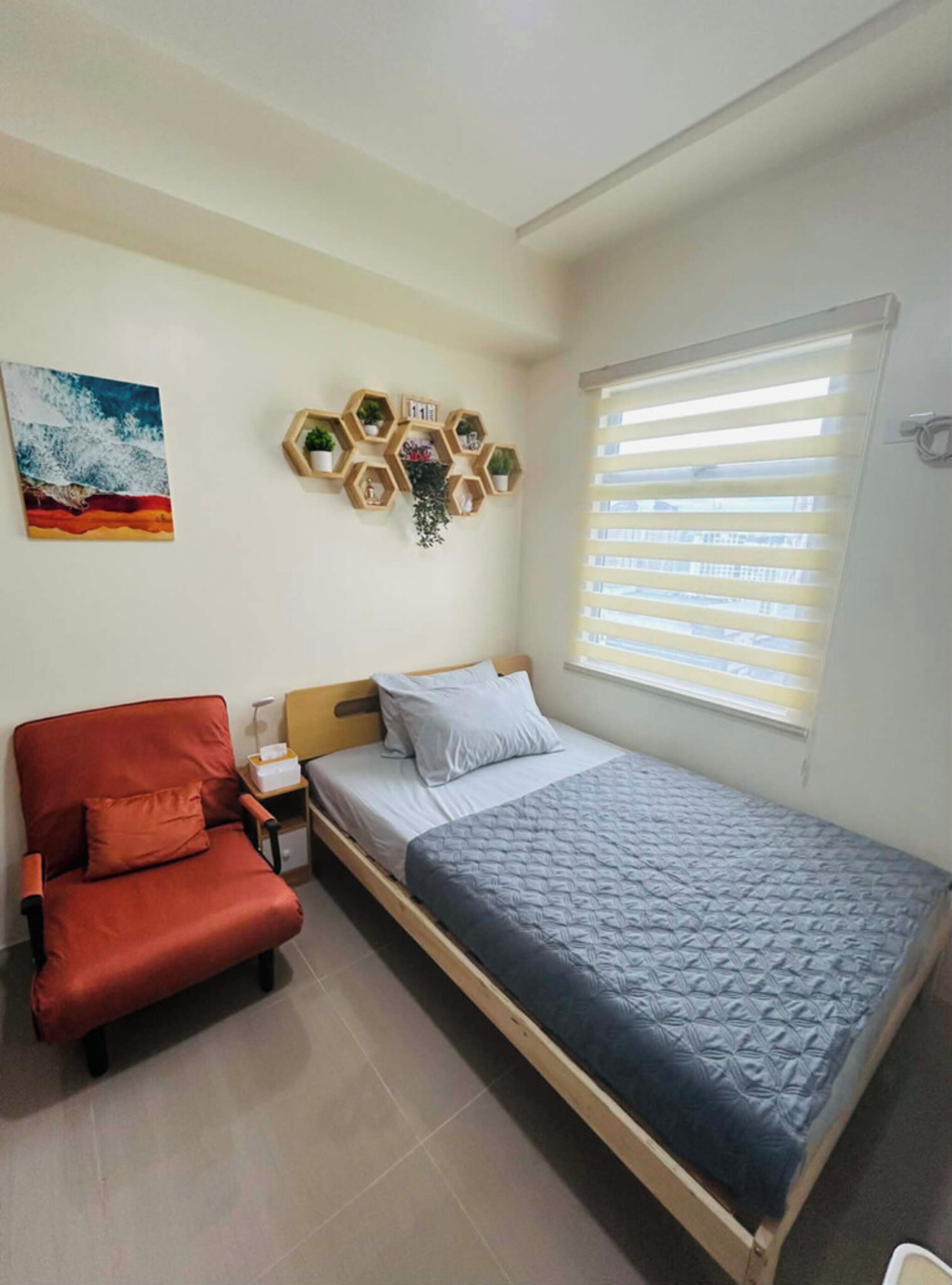 Une chambre à coucher comprenant un lit, une chaise, une étagère fixée au mur avec des plantes, un tableau et une fenêtre avec des stores.