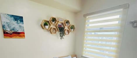 Um quarto com cama, uma cadeira, uma prateleira na parede com plantas, um quadro e uma janela com persianas.