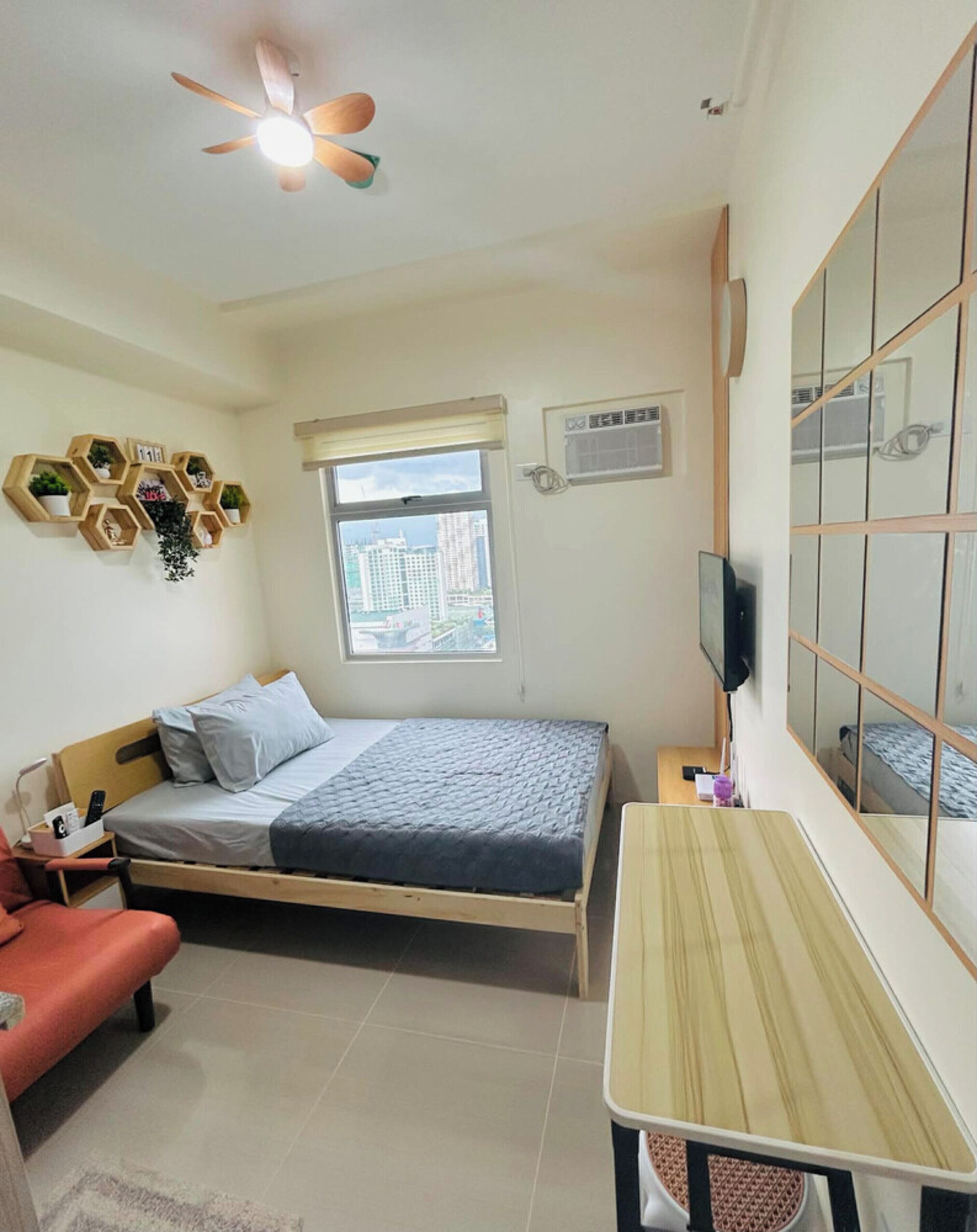Un dormitorio con cama, escritorio, silla, ventilador de techo, ventana y televisor montado en la pared.