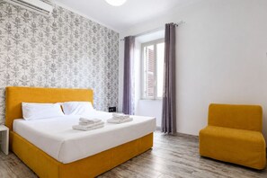 Quarto de hotel com uma cama grande, uma mesinha, uma janela com cortinas e uma poltrona amarela.