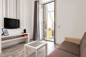 Een woonkamer met een flatscreentelevisie op een witte standaard, een kleine tafel, een bank en een schuifpui naar een buitenruimte.