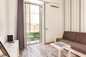 Ein Wohnzimmer mit einer Couch, einem Tisch, einer Schiebetür zum Balkon und einem Fernseher auf einem Schrank.
