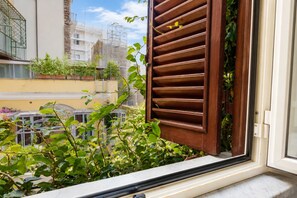 Ein Fenster mit Holzklappläden, die teilweise geöffnet sind, zeigt einen Blick auf einen Balkon mit Pflanzen und eine Stadt im Hintergrund.