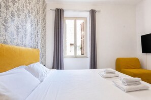 Ein Hotelzimmer mit Bett, einem Fenster mit Vorhängen, einem an der Wand befestigten Fernseher und einem Sessel.