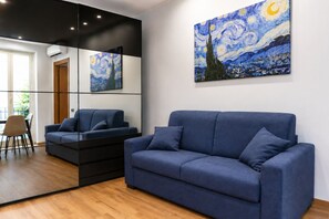 A modern living room with a blue sofa, a dining area with a table and chairs, and two paintings on the wall. - Elegantis Campo Marzio Penthouse With Terrace | Spanish Steps, Trevi & Pantheon (Roma)