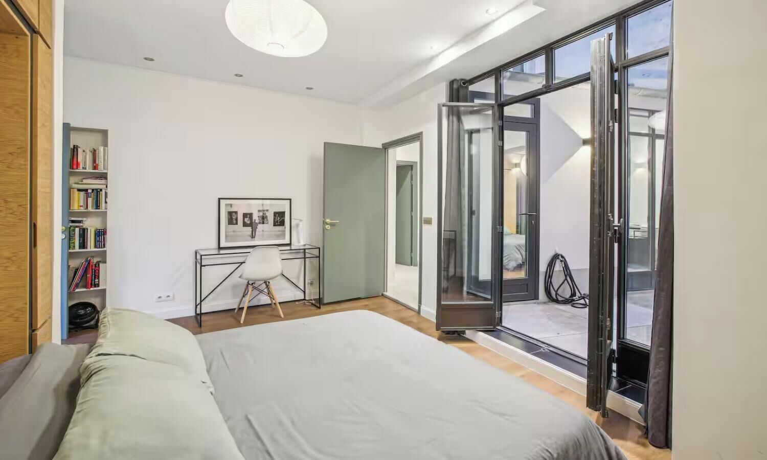 Un dormitorio con cama, escritorio, silla, estantería y puerta corrediza de cristal a un balcón.