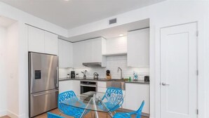 Une cuisine moderne avec des meubles blancs, des appareils en acier inoxydable et des chaises bleues disposées autour d’une table en verre.