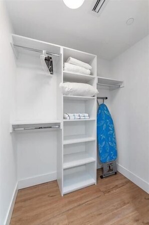 A white closet with shelves and hanging space, a blue ironing board, and a small appliance. - Deluxe 1BR near Bayfront Park &  Kaseya Center (Miami)