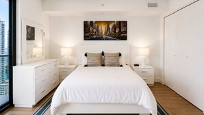 Un dormitorio moderno con una cama grande, lámparas de noche, un cuadro de paisaje urbano y vistas de edificios a través de una ventana.