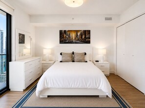 Ein modernes Schlafzimmer mit einem großen Bett, Nachttischlampen, einem Stadtbild und Blick auf Gebäude durch ein Fenster.
