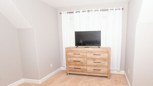A room with a beige carpet, a wooden dresser with drawers, a flat-screen TV on top, and white curtains. - SASADEERLODGE 6-bedroom house with AC in Richmond Hill (Richmond Hill)