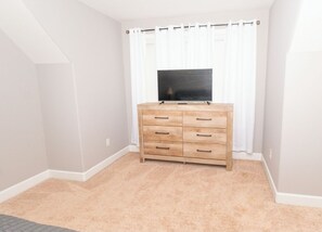 A room with a beige carpet, a wooden dresser with drawers, a flat-screen TV on top, and white curtains. - SASADEERLODGE 6-bedroom house with AC in Richmond Hill (Richmond Hill)