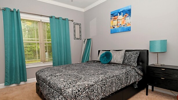 A bedroom with a bed, a nightstand, a lamp, a window with curtains, and a painting on the wall. - SASADEERLODGE 6-bedroom house with AC in Richmond Hill (Richmond Hill)