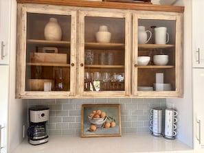 A kitchen with a wooden cabinet containing glassware and dishes, a coffee maker, and a framed painting of fruit. - Superb 3-bedroom home in peaceful Atascadero (Atascadero)