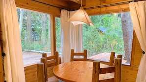 Un interior de madera con una mesa redonda y sillas, ventanas amplias con cortinas y una lámpara colgante.