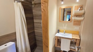 Un baño con suelo de madera, inodoro blanco, ducha con puerta de madera, lavabo con espejo arriba y un toallero.
