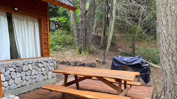 Una mesa de picnic de madera con bancos en una terraza, rodeada por un muro de piedra y un área boscosa.