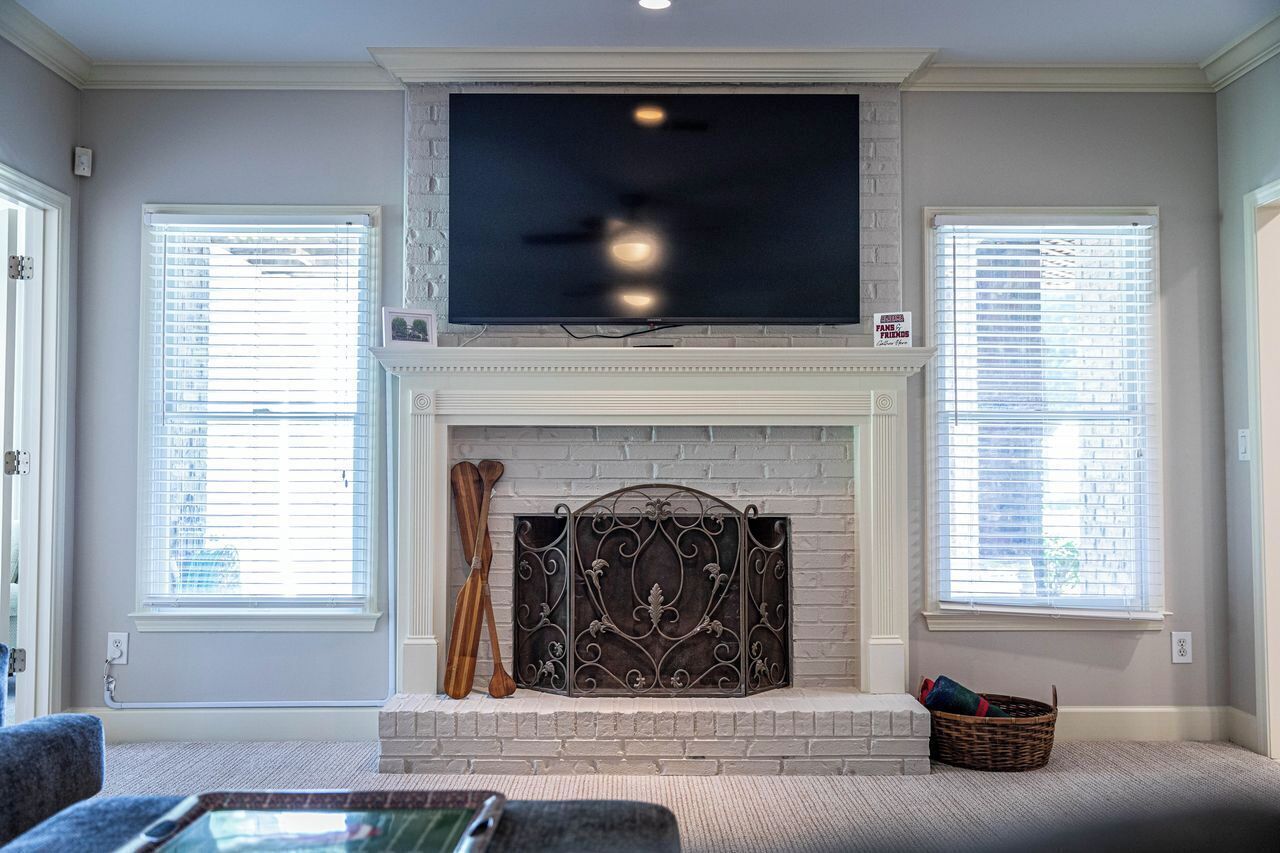 A living room with a fireplace, a television mounted above it, and two windows with blinds.