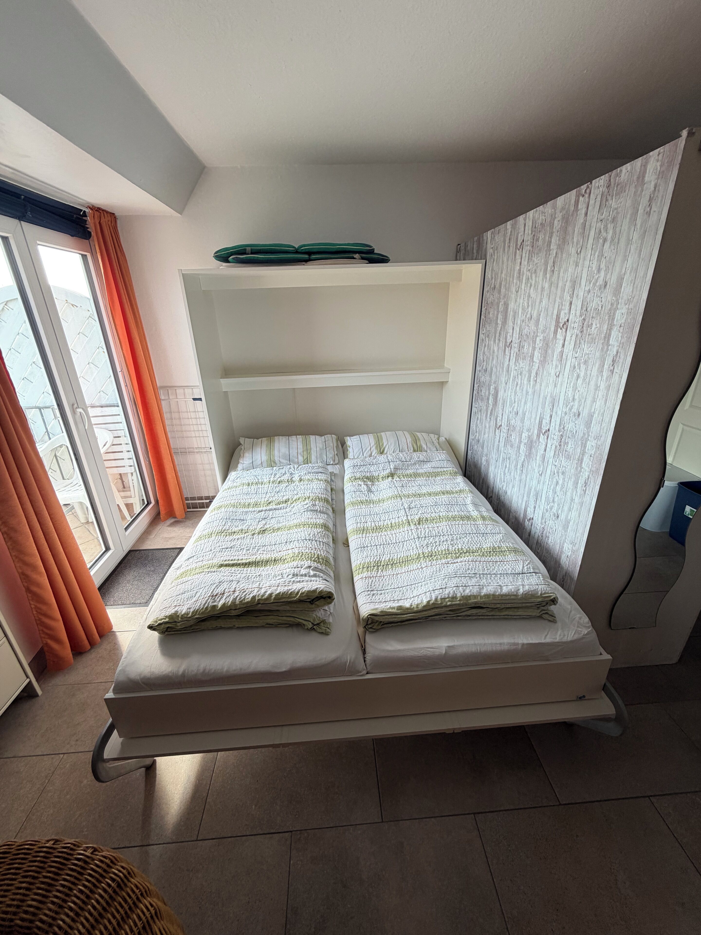 A single bed with a white headboard, a window with orange curtains, and a view of a building outside.