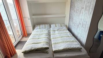 A single bed with a white headboard, a window with orange curtains, and a view of a building outside.